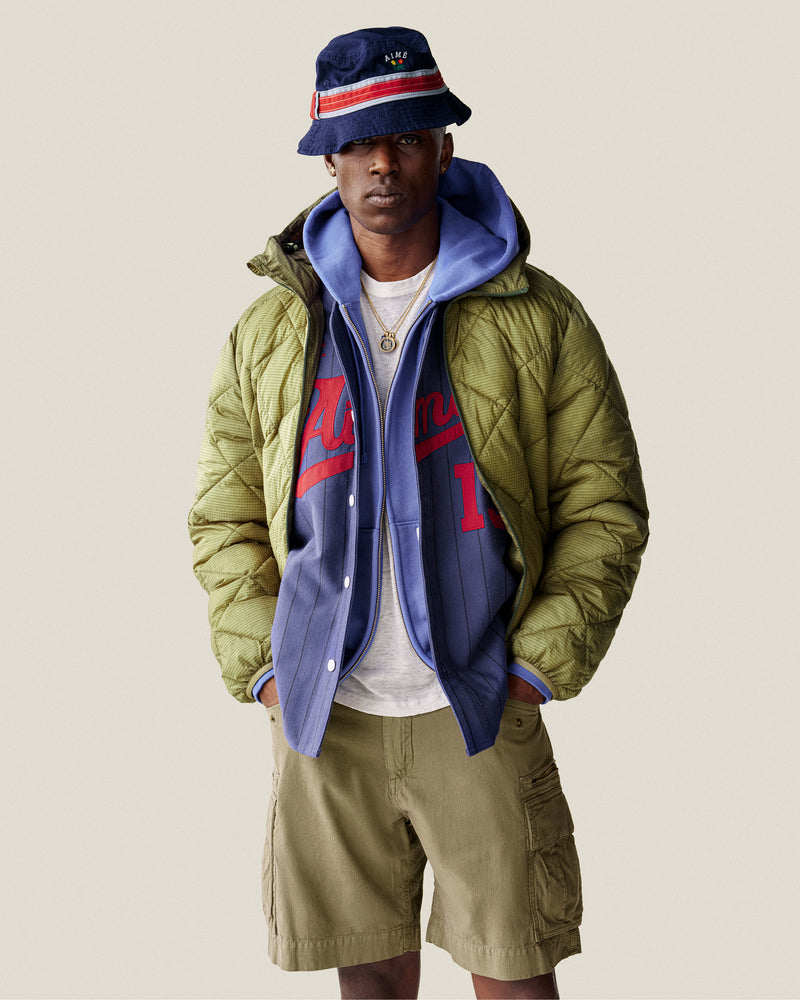 A man wears a blue bucket hat, blue hoodie, sports jersey, khaki cargo shorts, and a Nylon Quilted Unisphere Jacket.