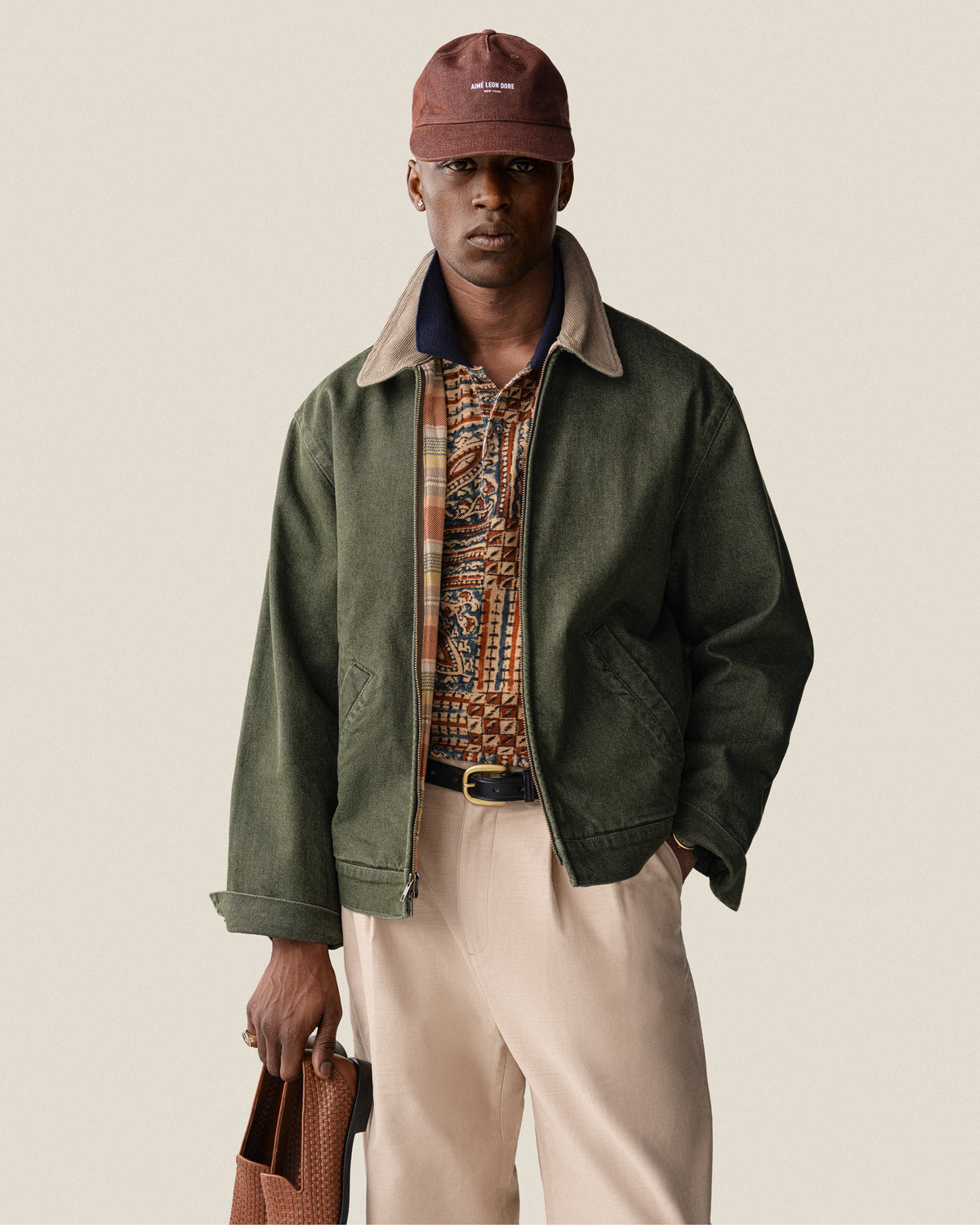 Man in green jacket, patterned shirt, beige pants, and Vintage Micro Logo Hat holding a brown bag against a plain background.