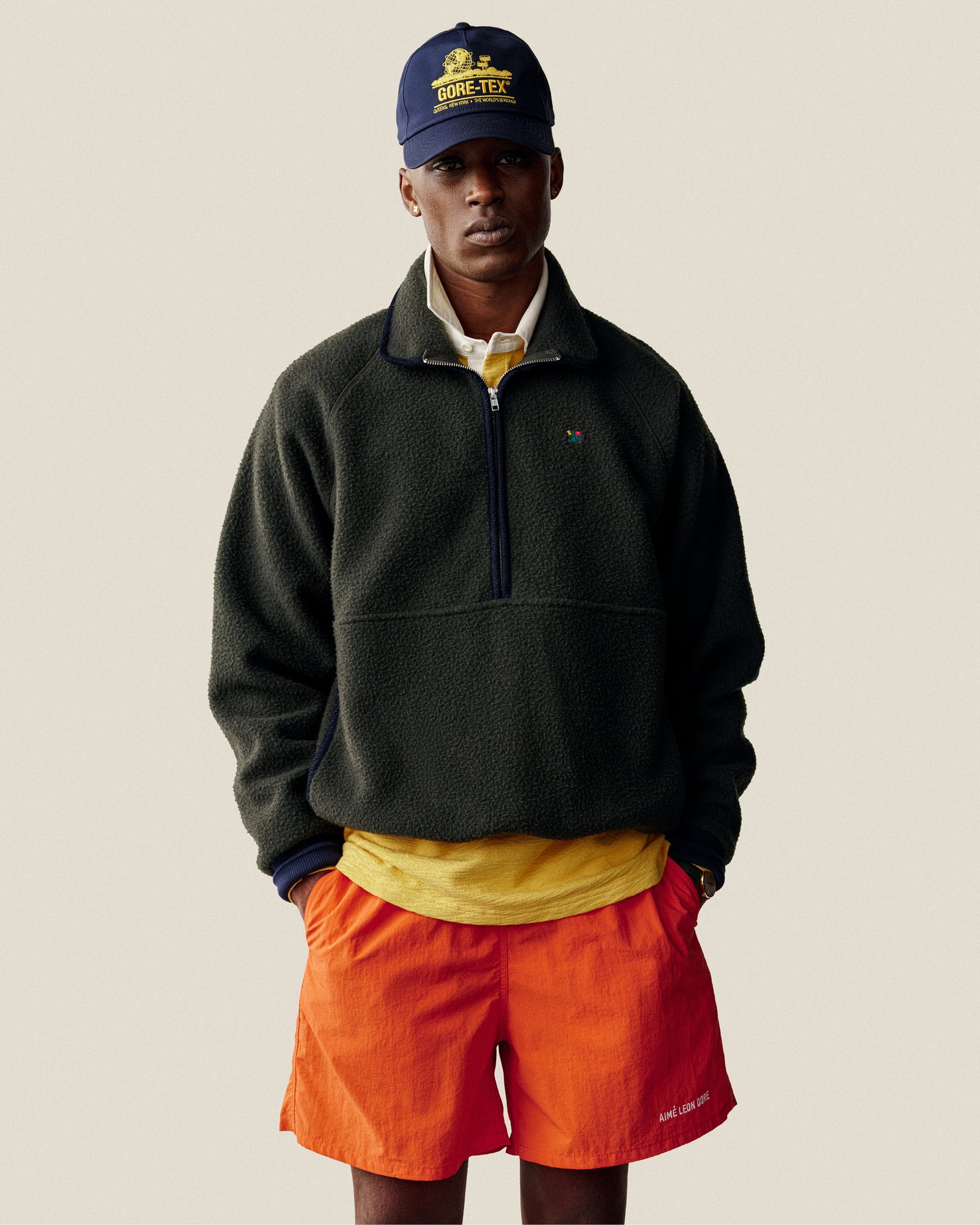 Man wearing a navy cap, yellow shirt, bright orange shorts, and Casentino Crest Half-Zip Pullover, hands in pockets.