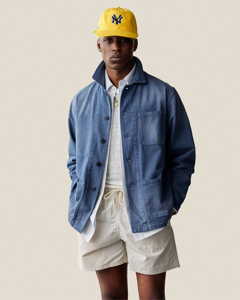 Man wearing an ALD / New Era Yankees Retro Fit Hat, blue jacket, white shirt, grey tee, beige shorts; plain background.