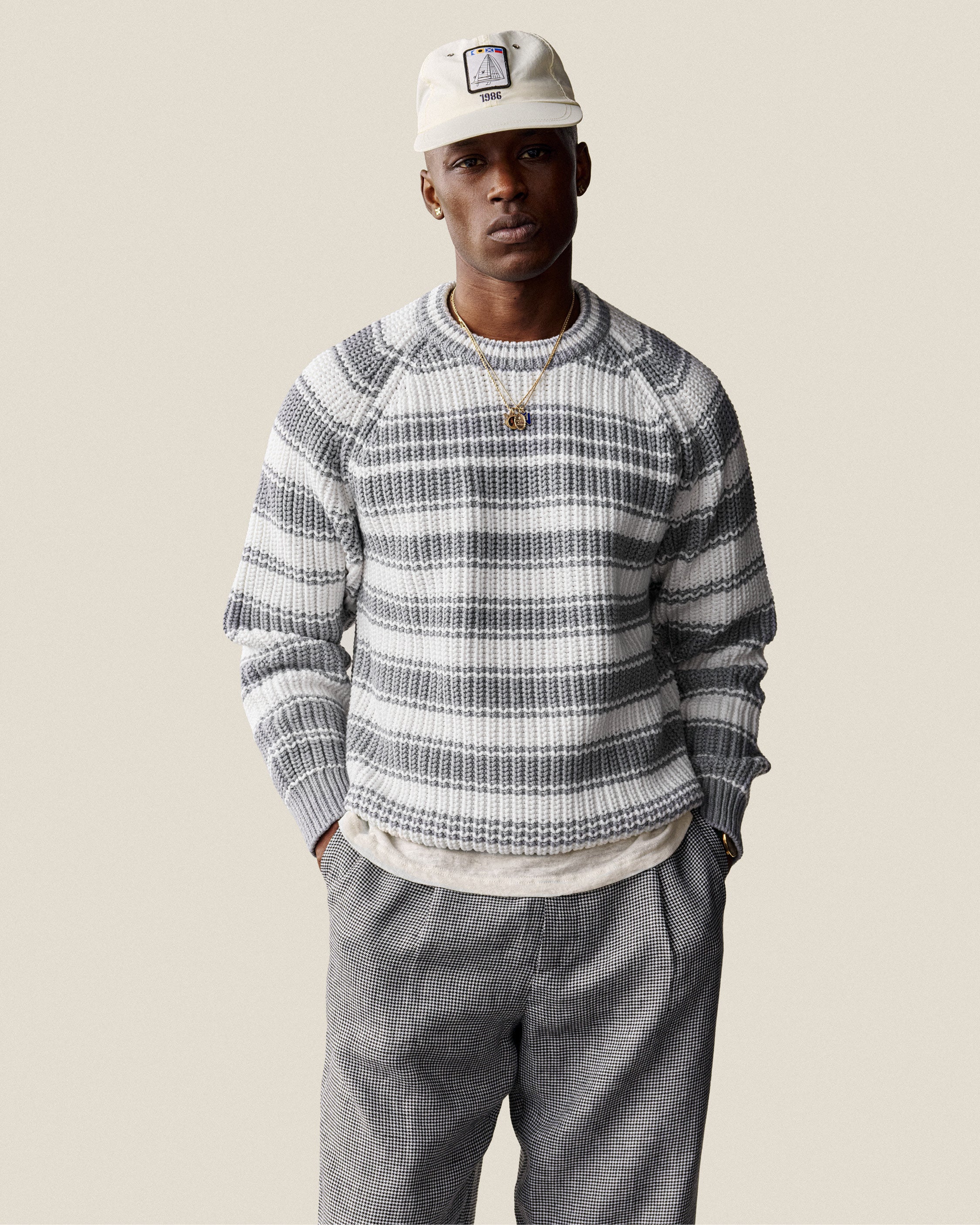 A man in a Striped Crewneck Sweater, gray pants, and a light cap stands with his hands in his pockets.