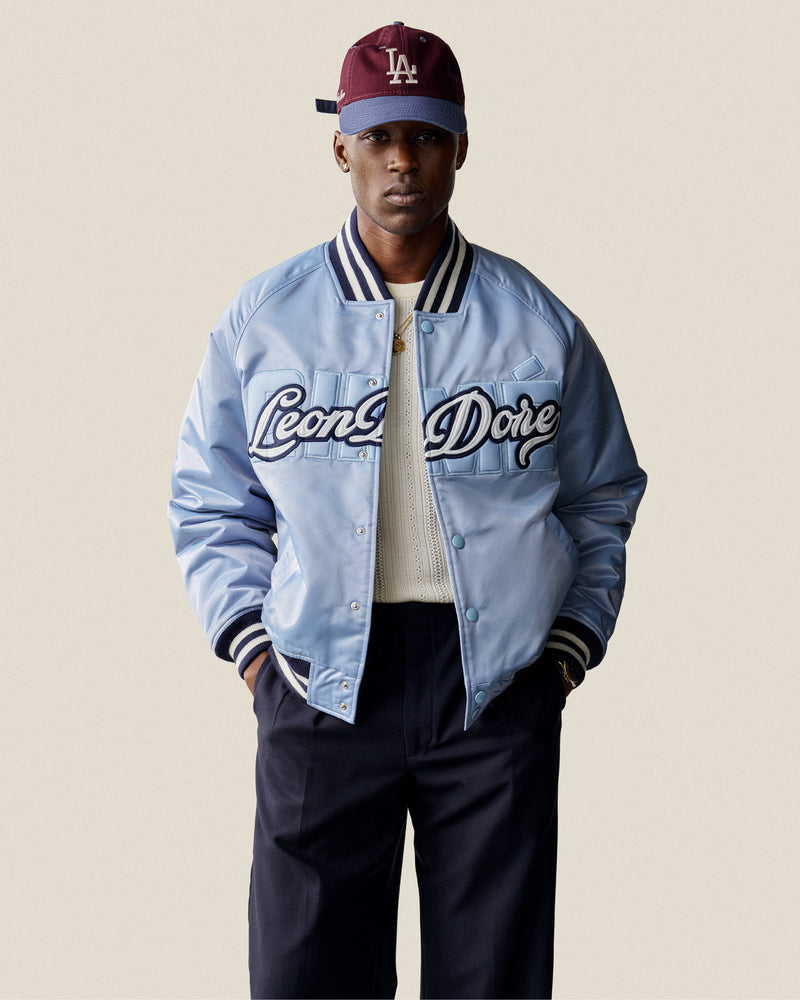 A man wears an Overlay Logo Satin Varsity Jacket, white sweater, navy pants, and a maroon LA cap, facing forward.