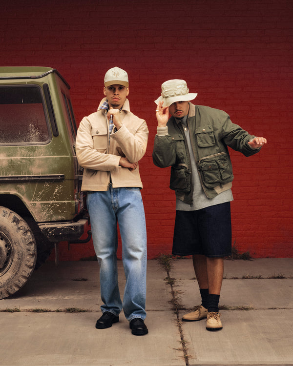 Two men pose by a green SUV; one wears jeans and a beige jacket, the other sports the Safari Utility Bomber.