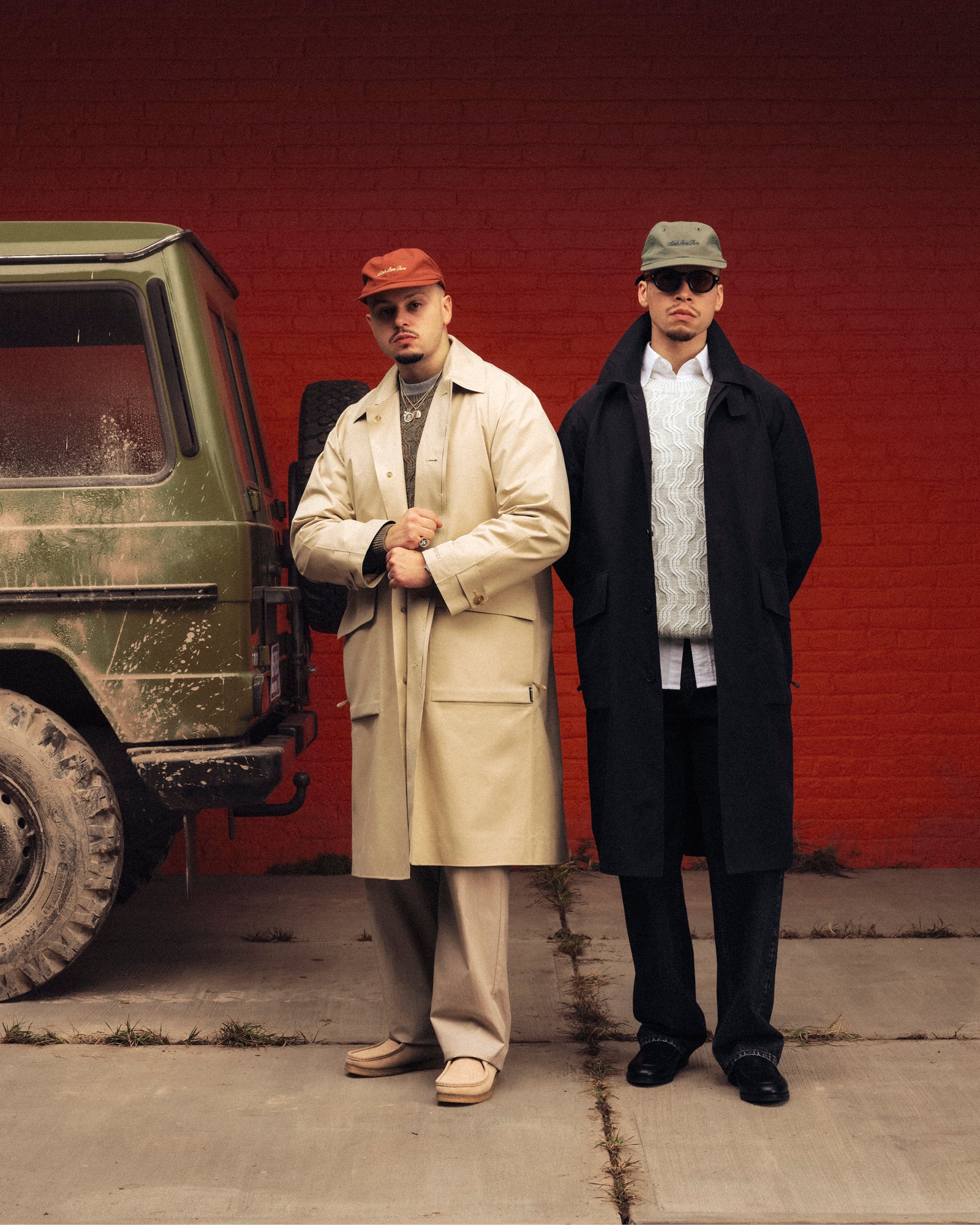 Two men in long coats wear Script Sport Hats and stand by a muddy SUV against a red brick wall, posing for the camera.