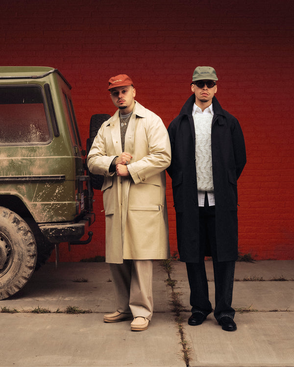 Two men in Linen Blend Cable Knit Sweaters stand by a muddy green SUV in front of a red brick wall.