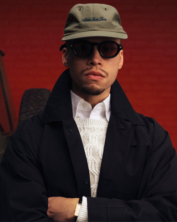 Man wearing a Script Sport Hat, black sunglasses, black coat, and white sweater stands by a red brick wall.