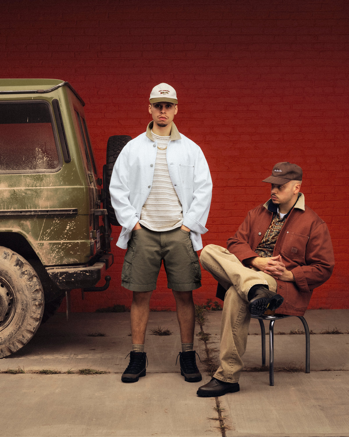 Two men in casual outfits, one wearing the Colorblock Lure Hat, pose by a muddy SUV and red brick wall.