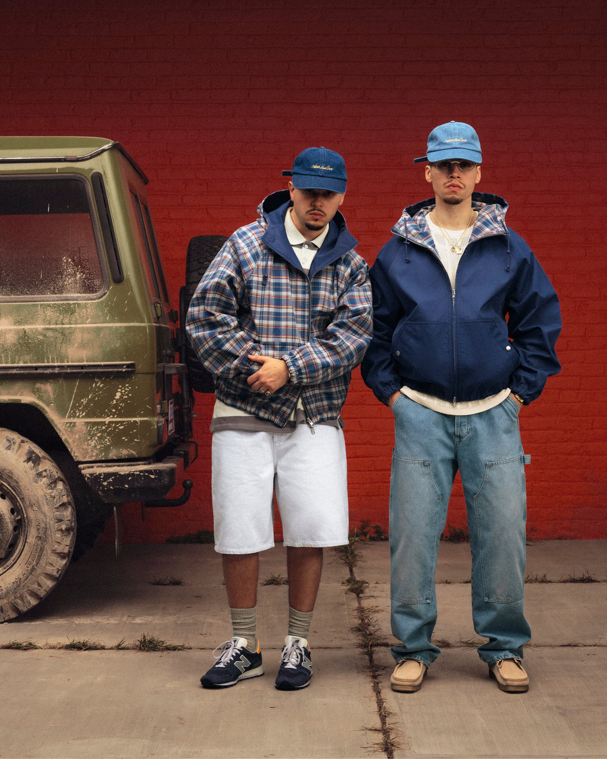 Two men in Reversible Madras Windbreakers and blue caps stand by a muddy green SUV in front of a red brick wall.