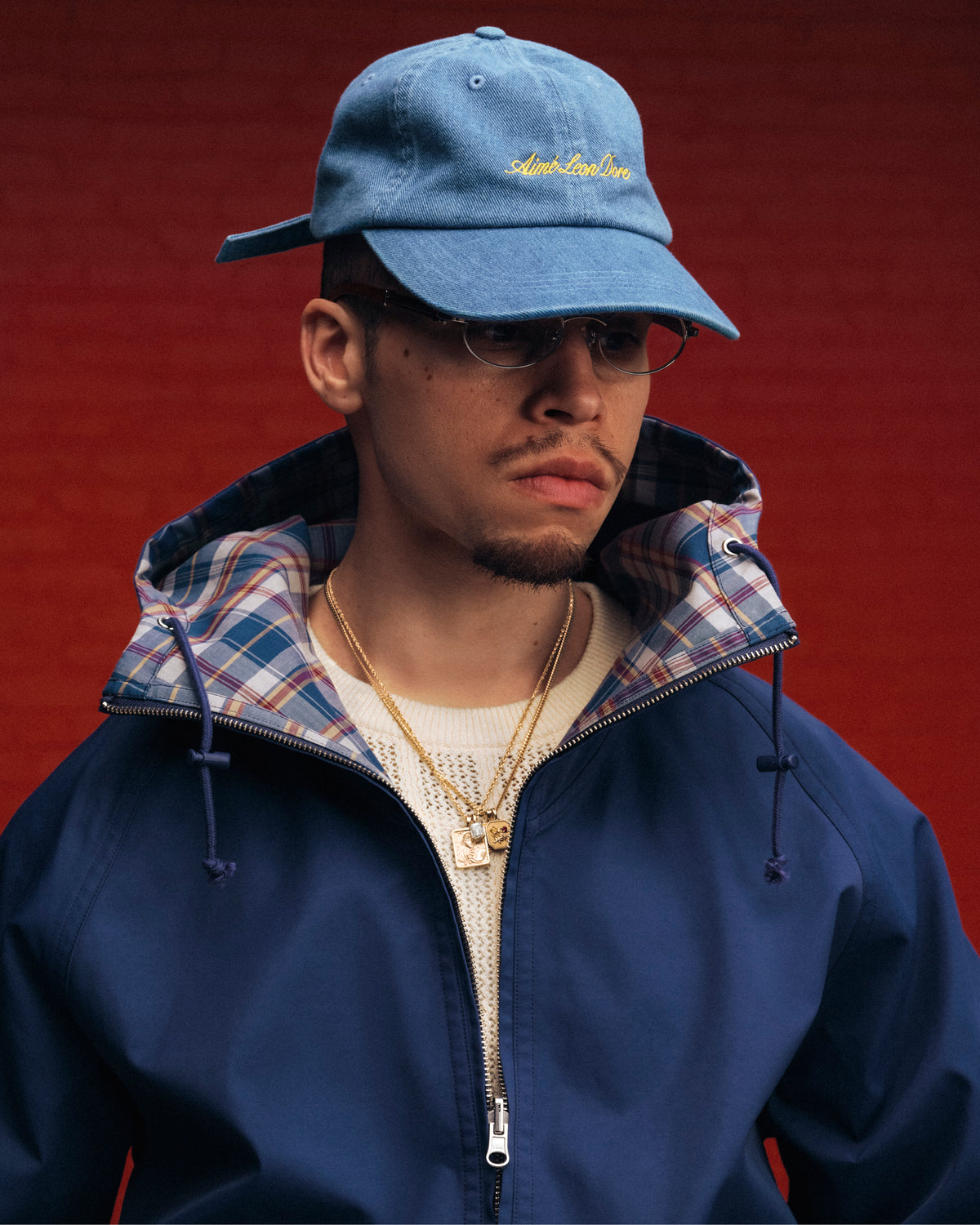A man in glasses, gold necklaces, and a blue Denim Script Hat stands against a red background.