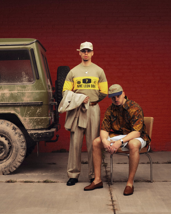 Two men pose by a muddy SUV; one stands in beige trousers and a Safari Heritage Hat, the other sits in shorts.