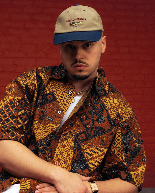 A man in a patterned shirt and the Colorblock Lure Hat poses against a red brick wall, looking at the camera.