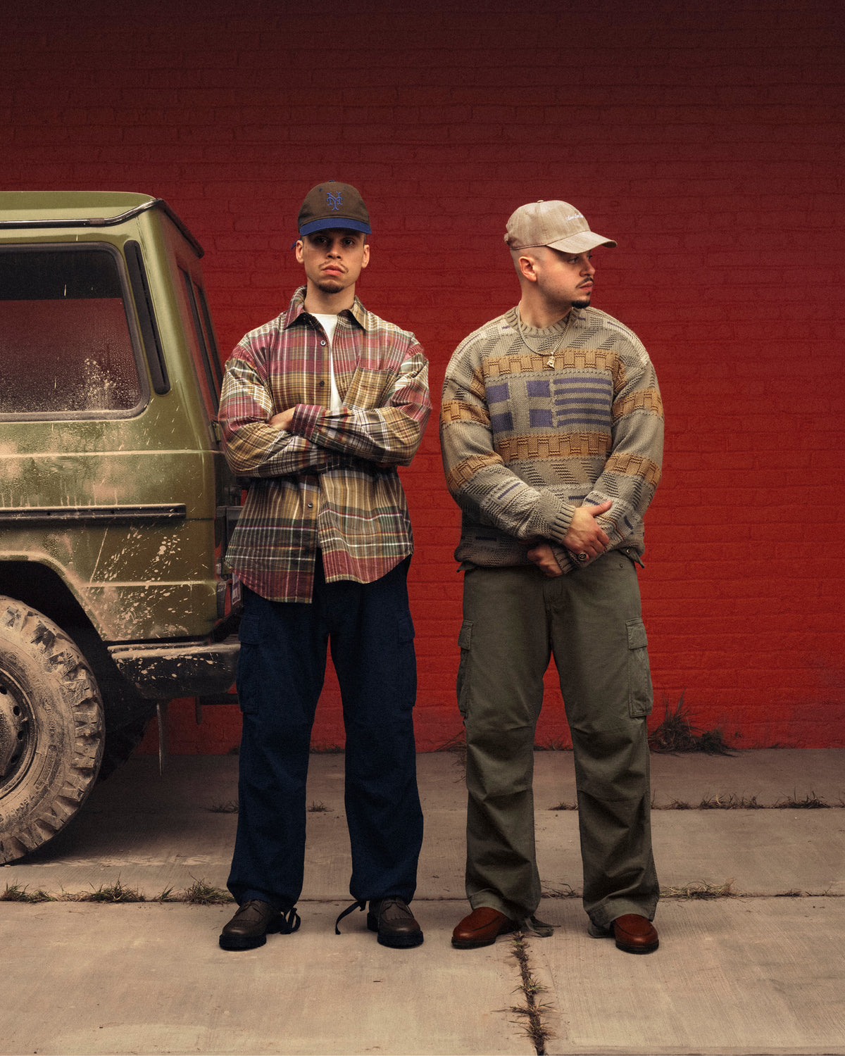 Two men in Washed Madras Shirts stand by a green SUV and a red brick wall, both looking serious.