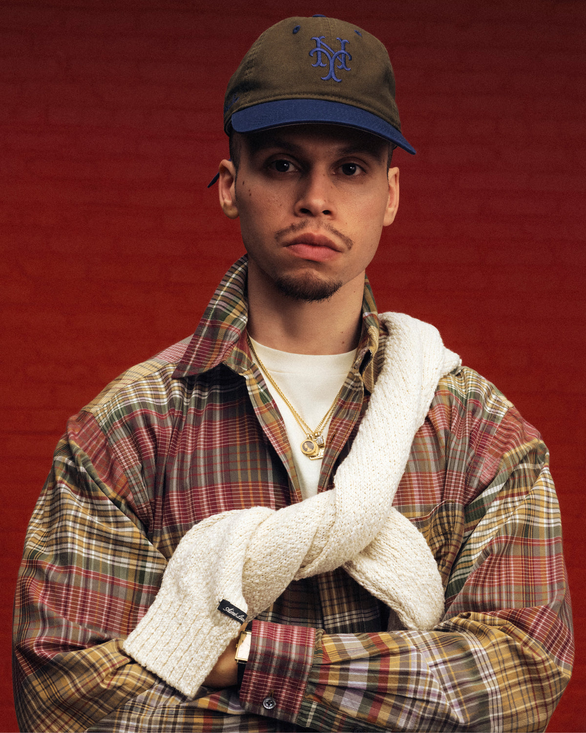 A man in a plaid shirt, gold chains, and an ALD / New York Mets Colorblock Ballpark Hat stands with a sweater over his shoulders.