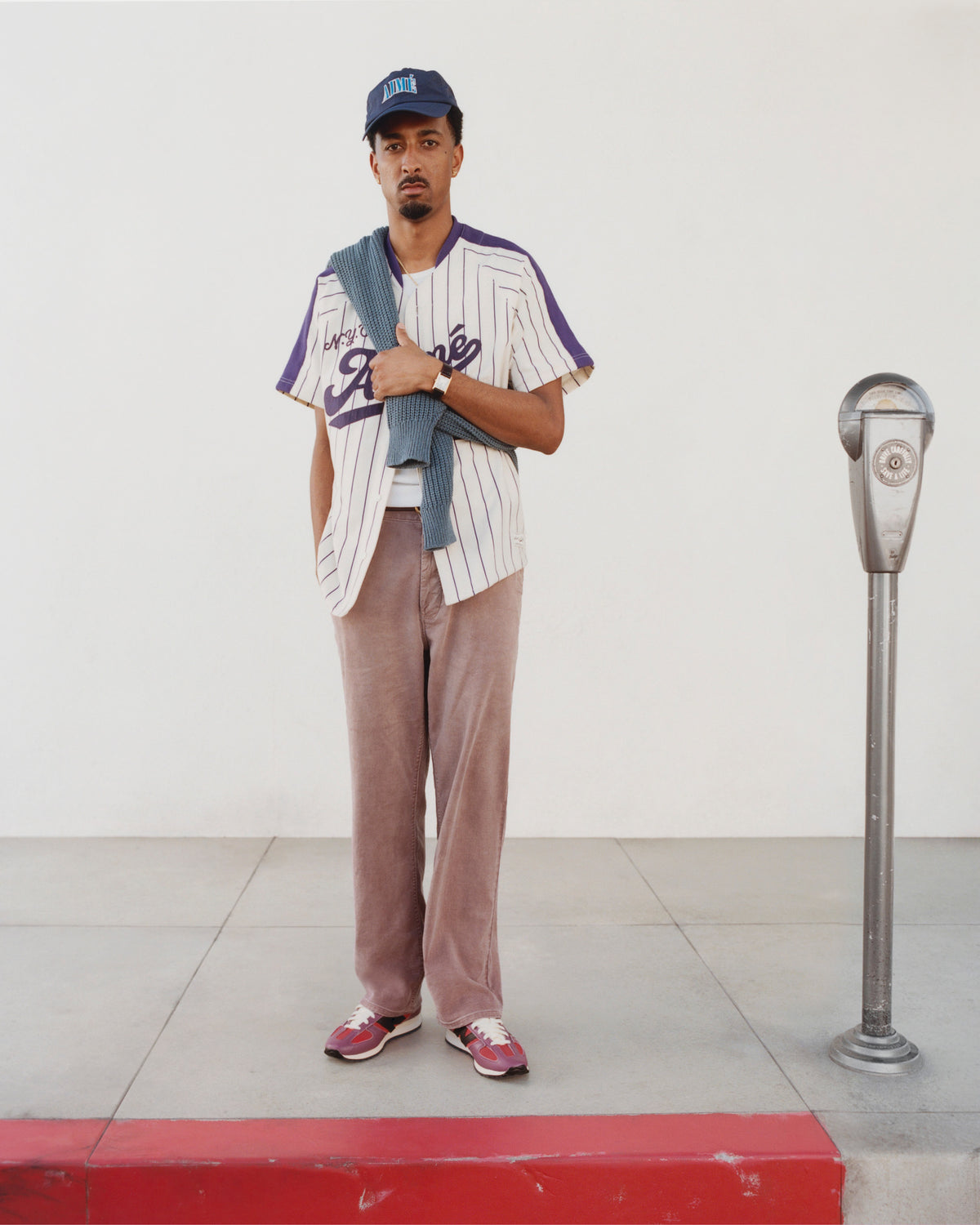 Man stands by a parking meter wearing Linen Blend Carpenter Pant, baseball jersey, and cap, sweater on his shoulder.