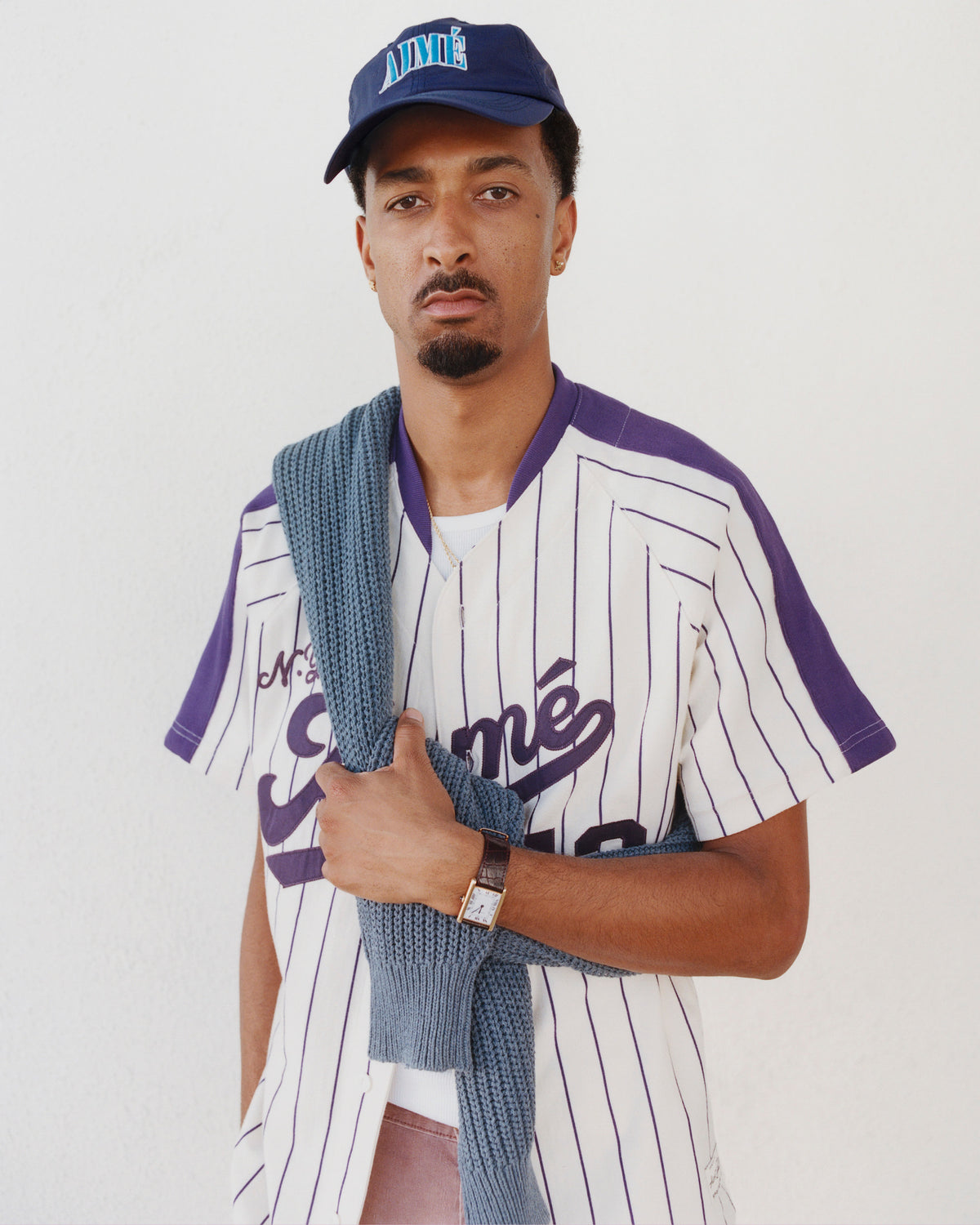 Man in a striped baseball jersey poses against a white wall, wearing a blue Nylon Crusher Hat and sweater over his shoulder.