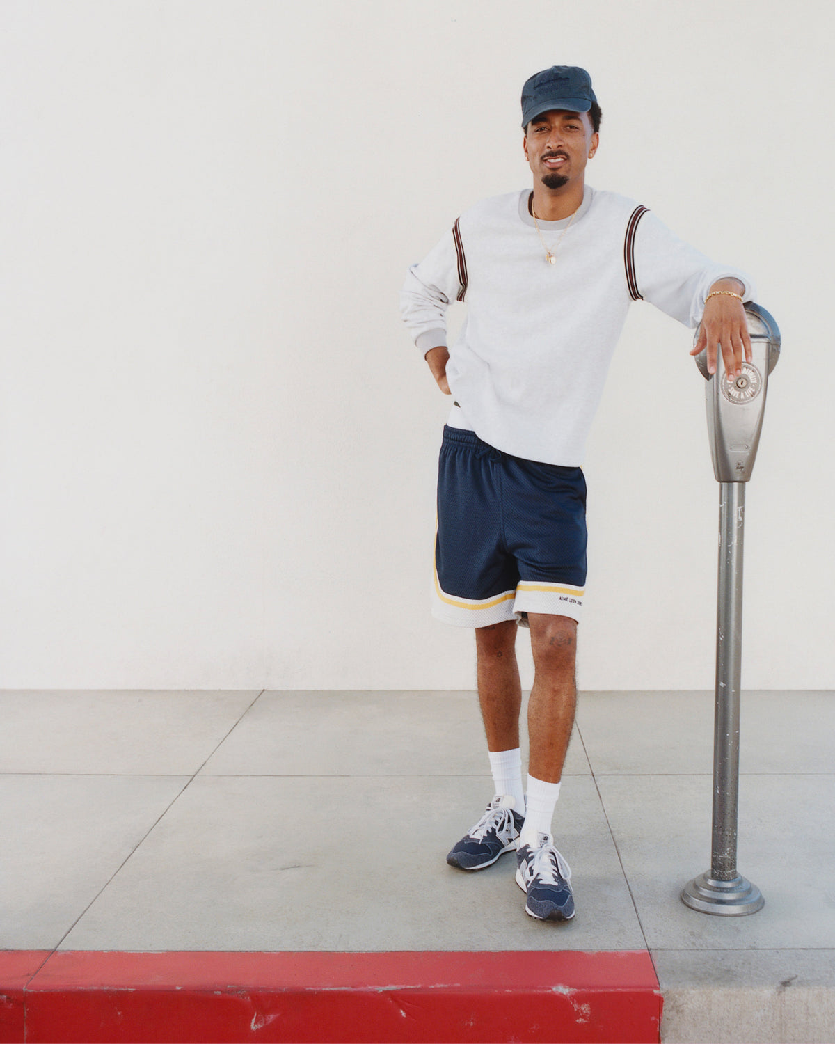 Man in a white sweatshirt, navy shorts, and Sun Faded Unisphere Hat leans on a parking meter by a white wall.