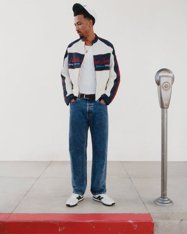 Man in a Nylon Moto Jacket, jeans, and white sneakers stands by a parking meter on the sidewalk, looking down.