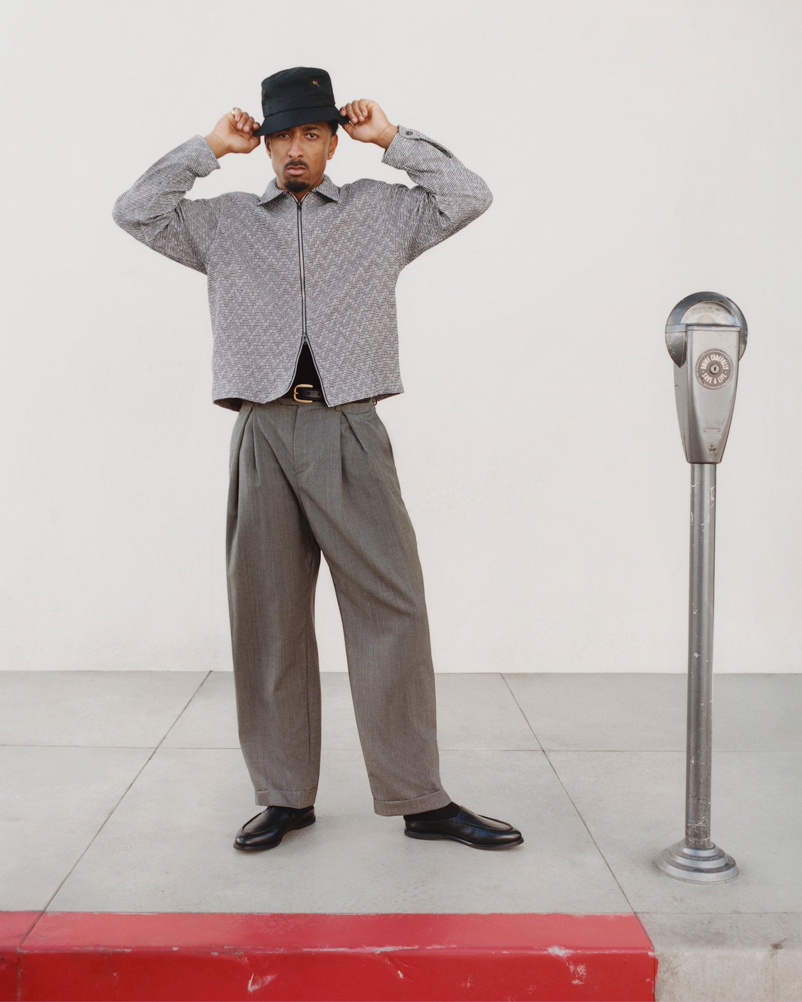 Man in Textured Zip Shirt and wide trousers adjusts black bucket hat beside a parking meter on the sidewalk.