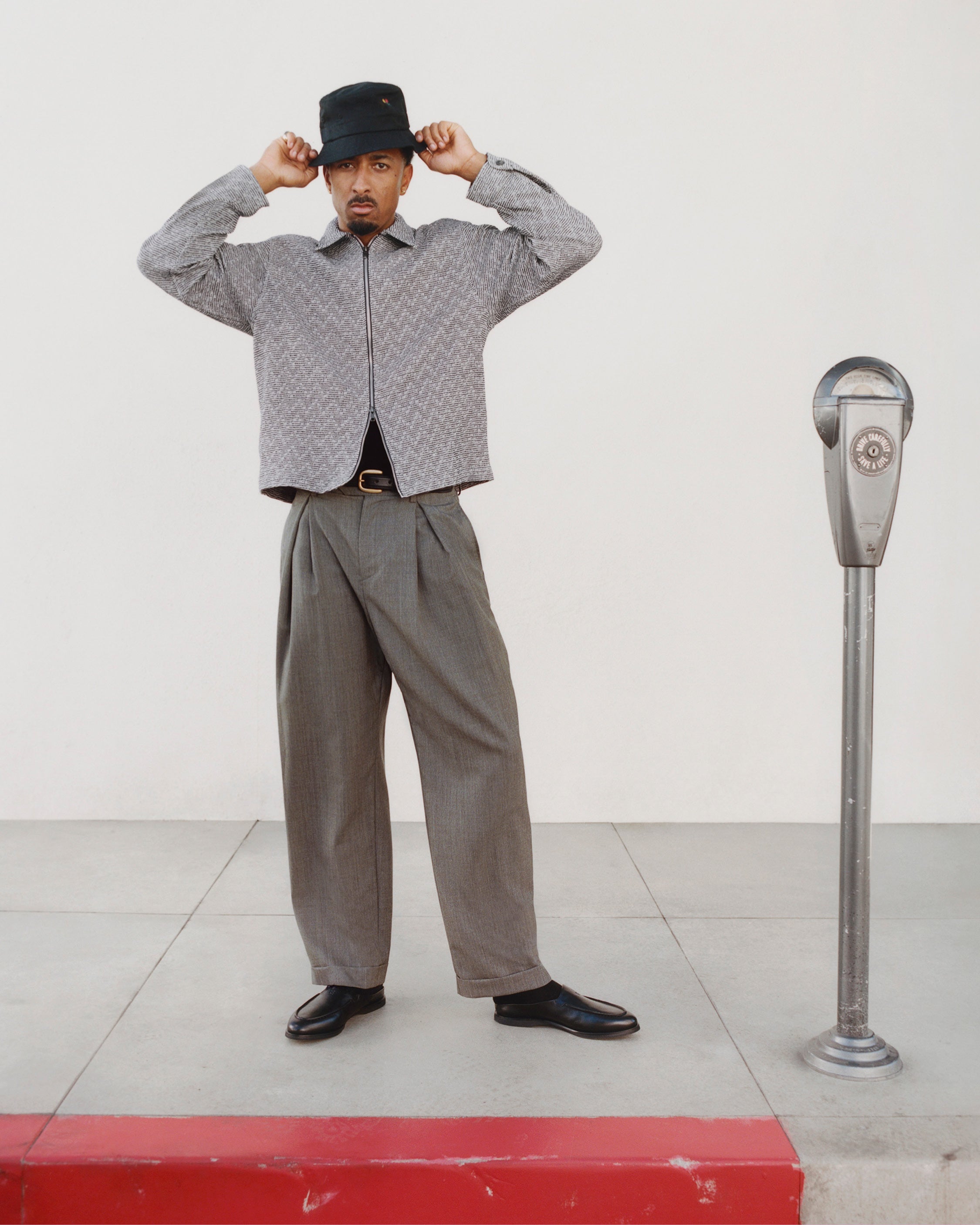 Man in Textured Zip Shirt and wide trousers adjusts black bucket hat beside a parking meter on the sidewalk.