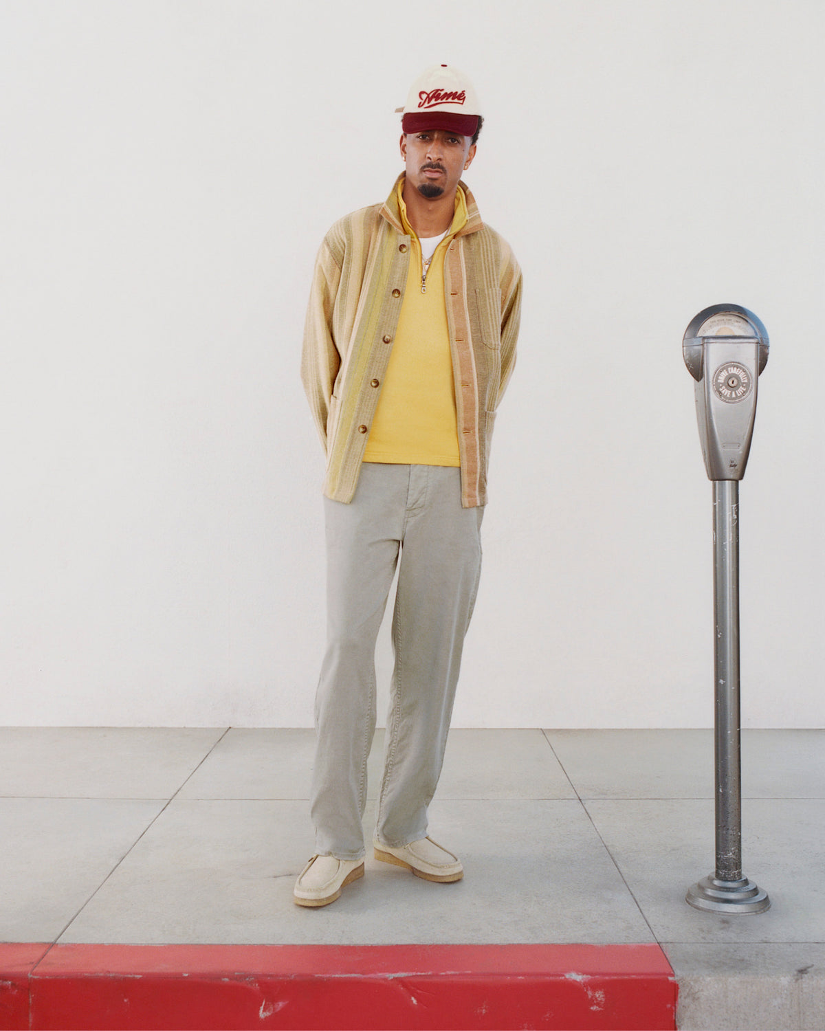 Man wears Linen Blend Carpenter Pant, yellow shirt, and red cap, standing by a parking meter against a white wall.
