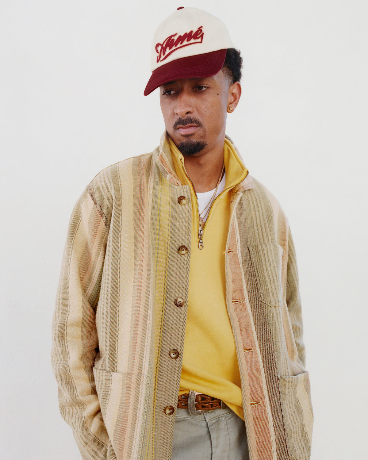 Man wearing a striped jacket, yellow shirt, and the Cycling Logo Hat, looking down with hands in jacket pockets.
