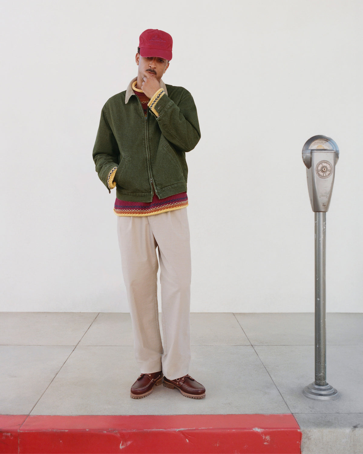 Man in Sun Faded Unisphere Hat, green jacket, beige pants, brown shoes stands by a parking meter on sidewalk.