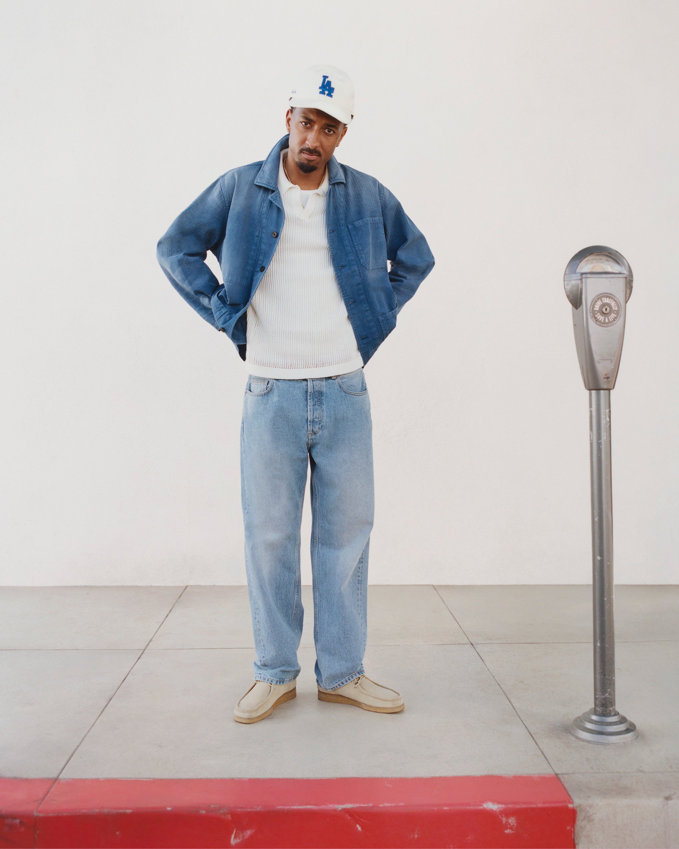 Man in Sun Faded Chore Jacket, jeans, white shirt and cap stands by parking meter on sidewalk with red curb.