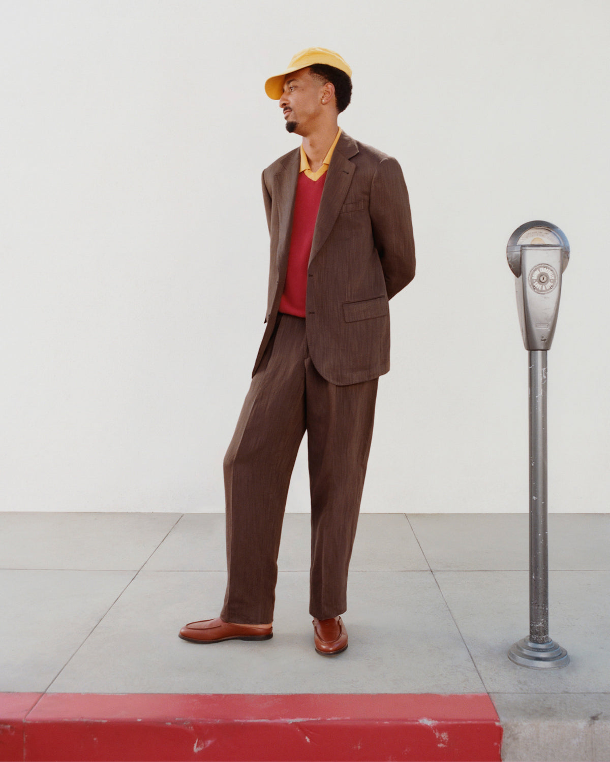 A man in Linen Blend Suit Trouser and a yellow cap stands by a parking meter with hands behind his back on the sidewalk.