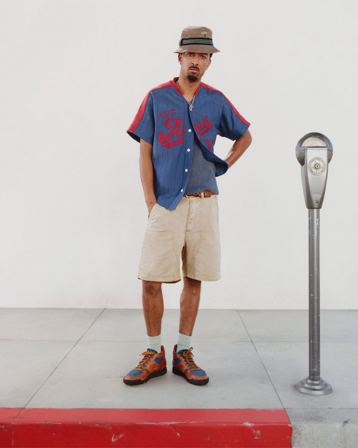 A man in an Aimé Baseball Jersey stands on a sidewalk next to a parking meter and red curb.