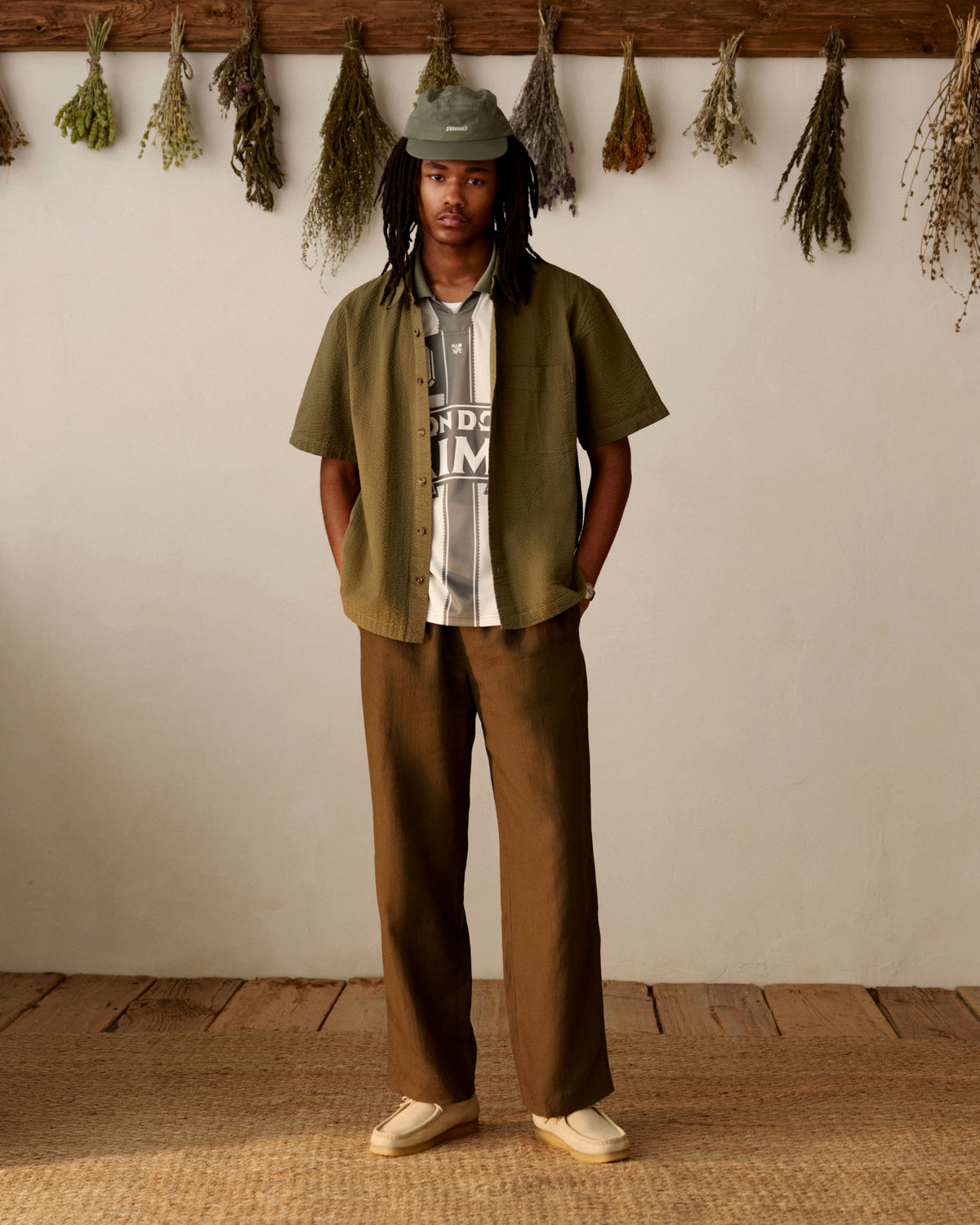 Person in olive shirt, striped shirt, Linen Leisure Pant, and loafers stands indoors by dried hanging herbs.