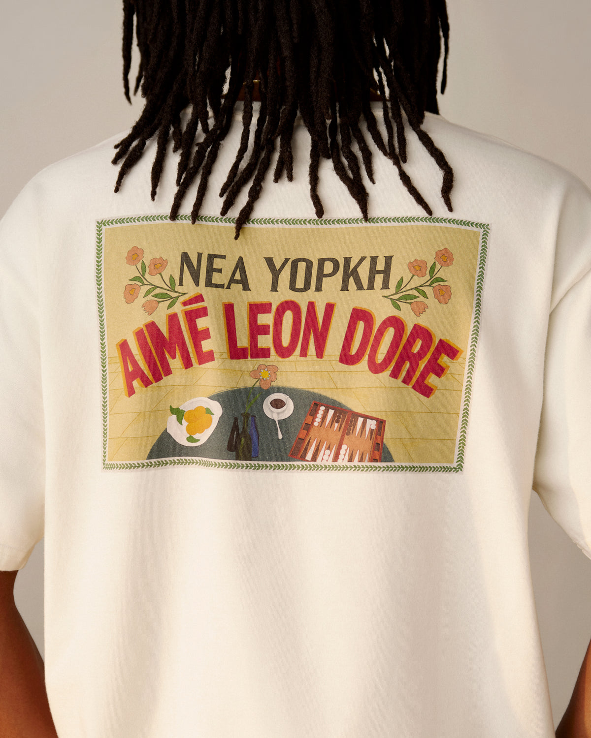 Person wearing a white Aimé Postcard Tee with NEA YOPKH AIMÉ LEON DORE and graphic art on the back.
