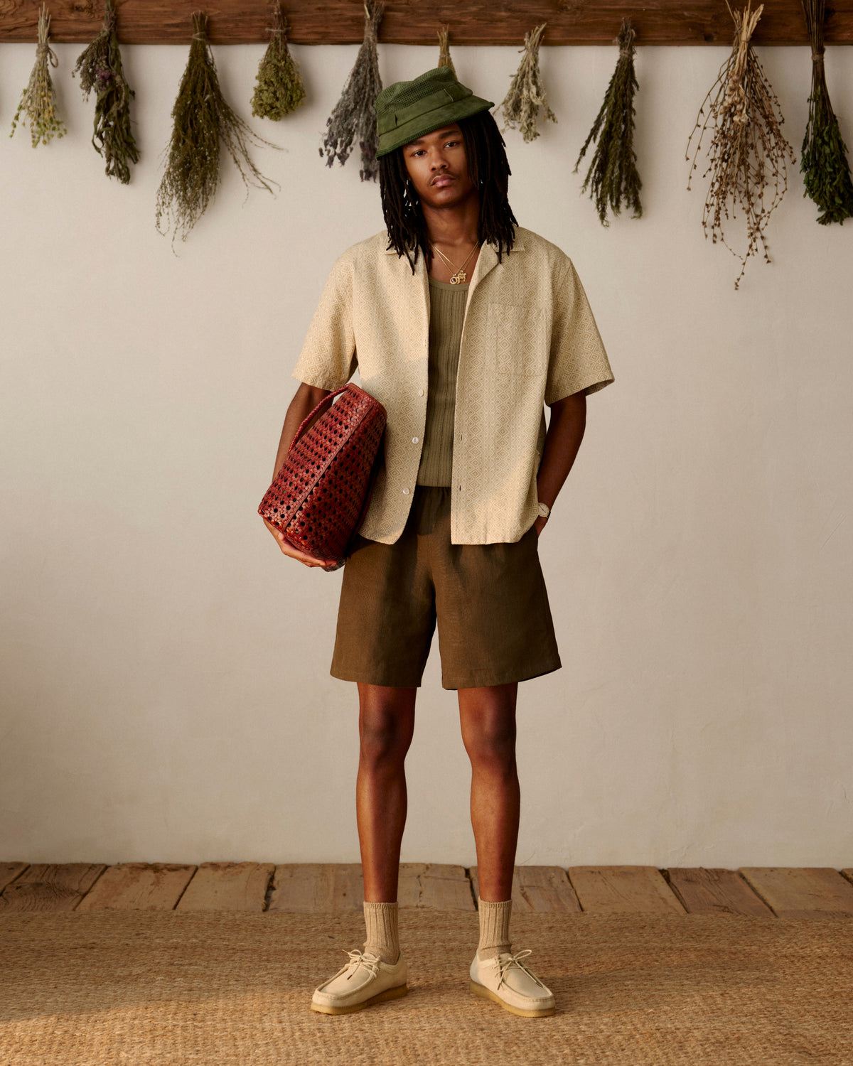 A person in a beige shirt and brown shorts holds a Woven Leather Tote Bag, with dried herbs hanging behind.