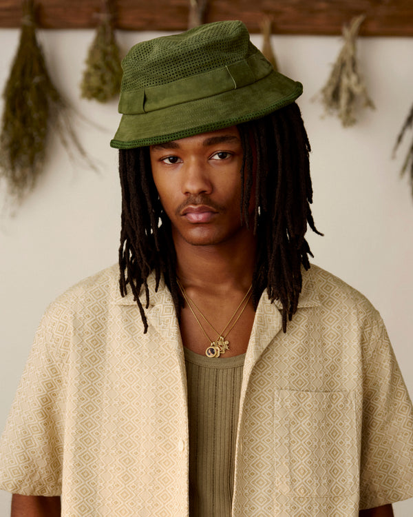 Young person with long dreadlocks wears a Perforated Suede Bucket Hat, beige shirt, and layered necklaces indoors.