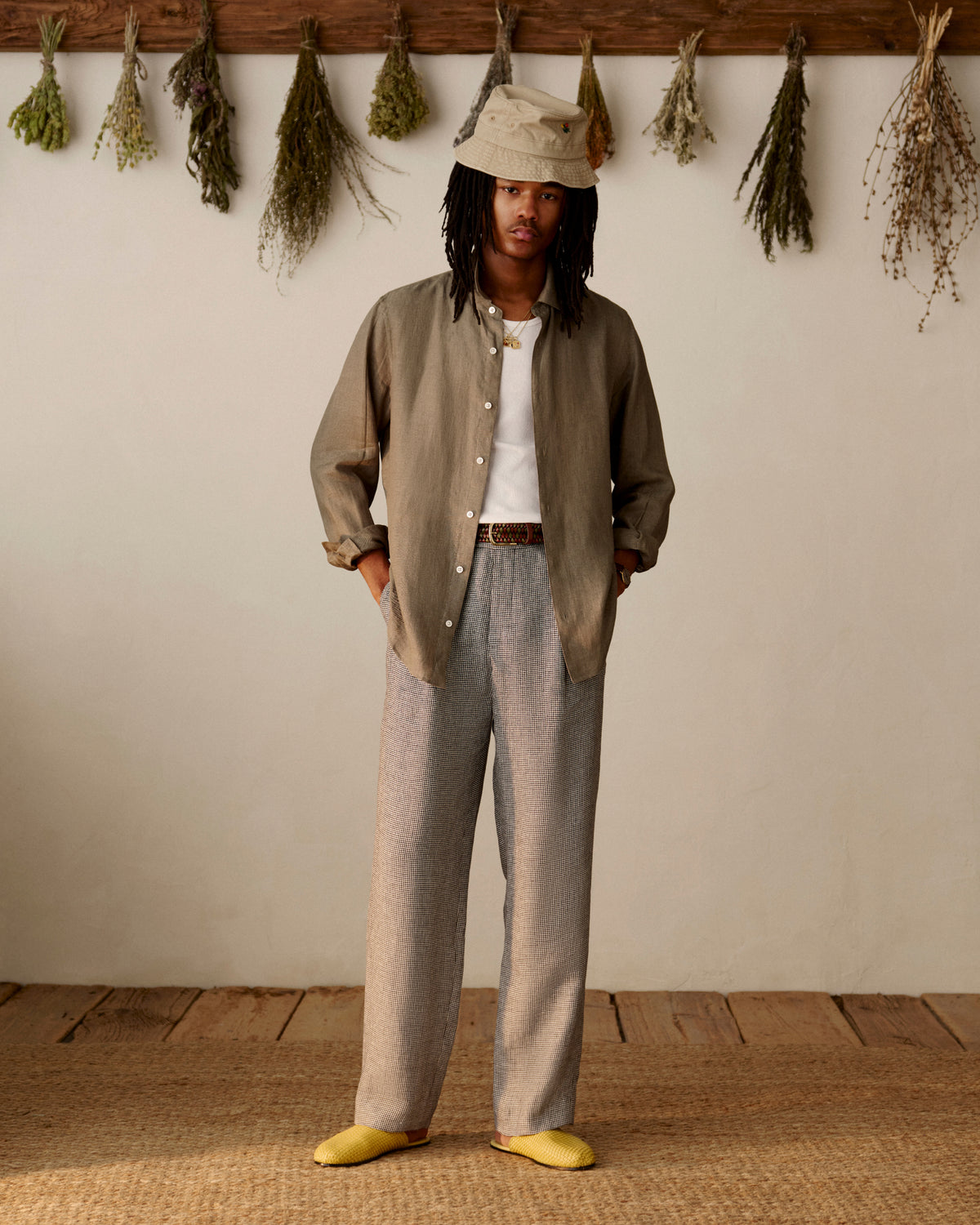 A person in Houndstooth Pleated Trousers and a bucket hat stands indoors with dried herbs hanging on the wall behind them.