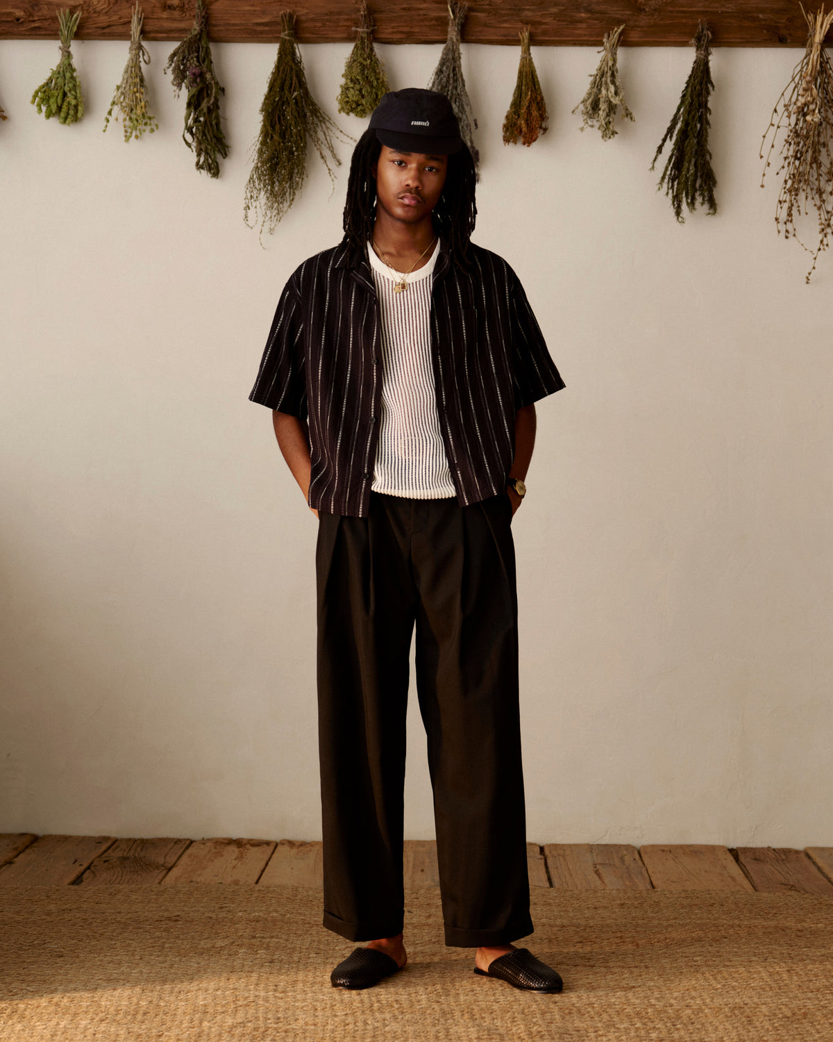 A man in a Cropped Jacquard Camp Shirt and loose black pants stands indoors with dried herbs hanging on the wall behind him.