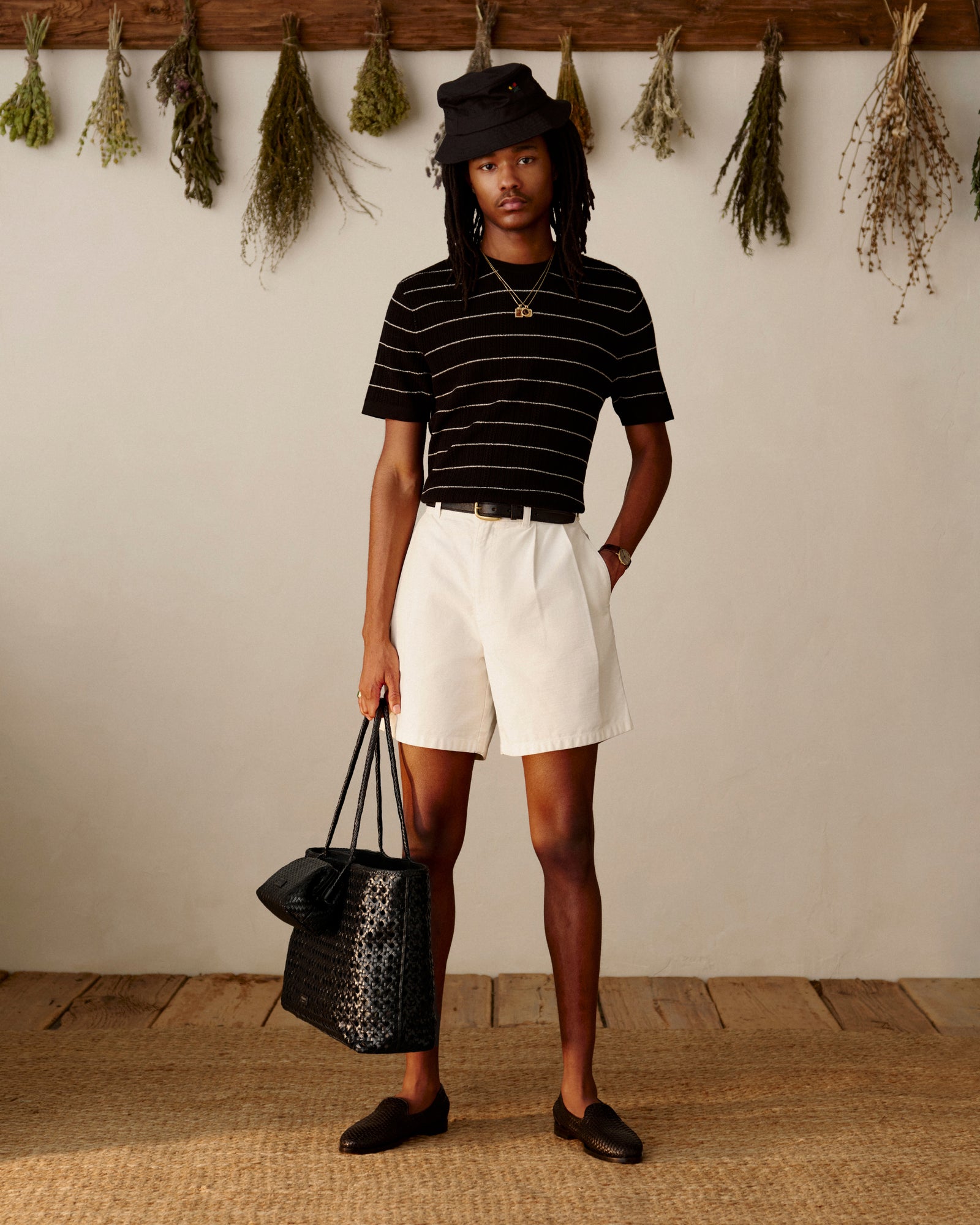 Person in a black hat, striped shirt, white shorts, loafers holds a Woven Leather Shoulder Bag; herbs hang behind.