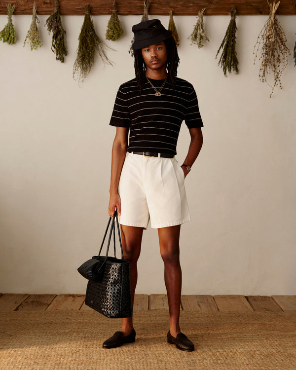 A person wears the Merino Wool Eyelet Tee, white shorts, black hat and loafers, holding a large black bag indoors.