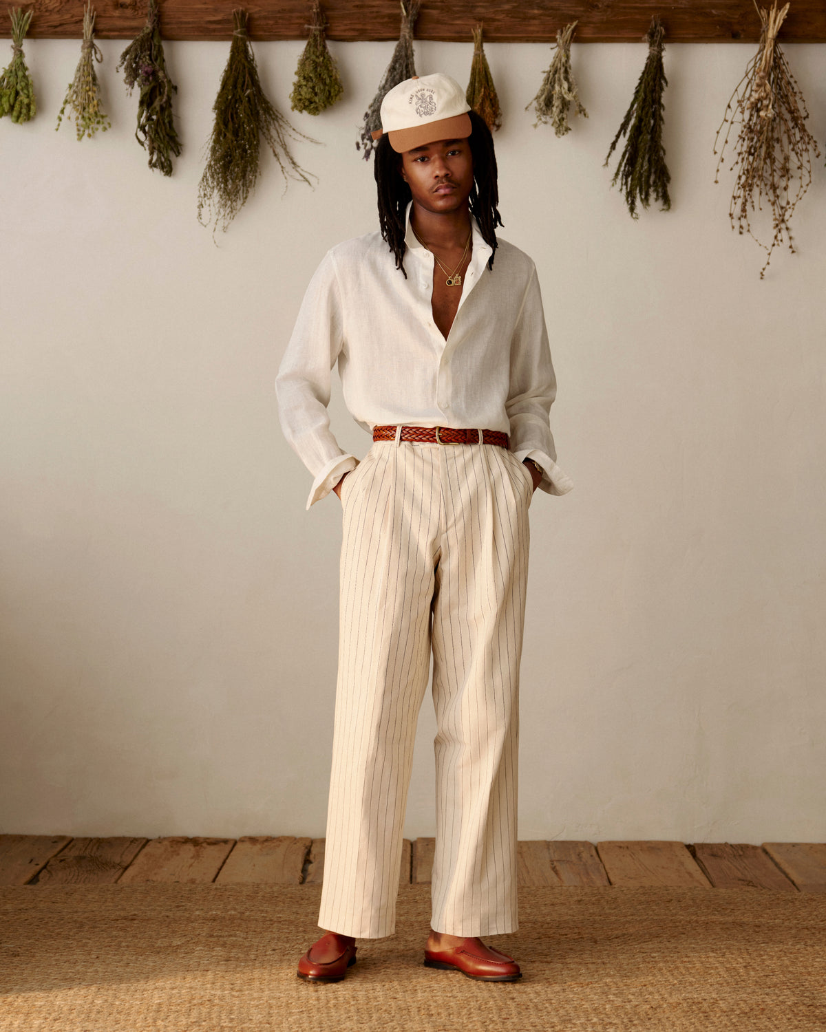 A person in a cream shirt, Double Pleated Pinstripe Suit Trouser, loafers, and a cap stands indoors near dried herbs.