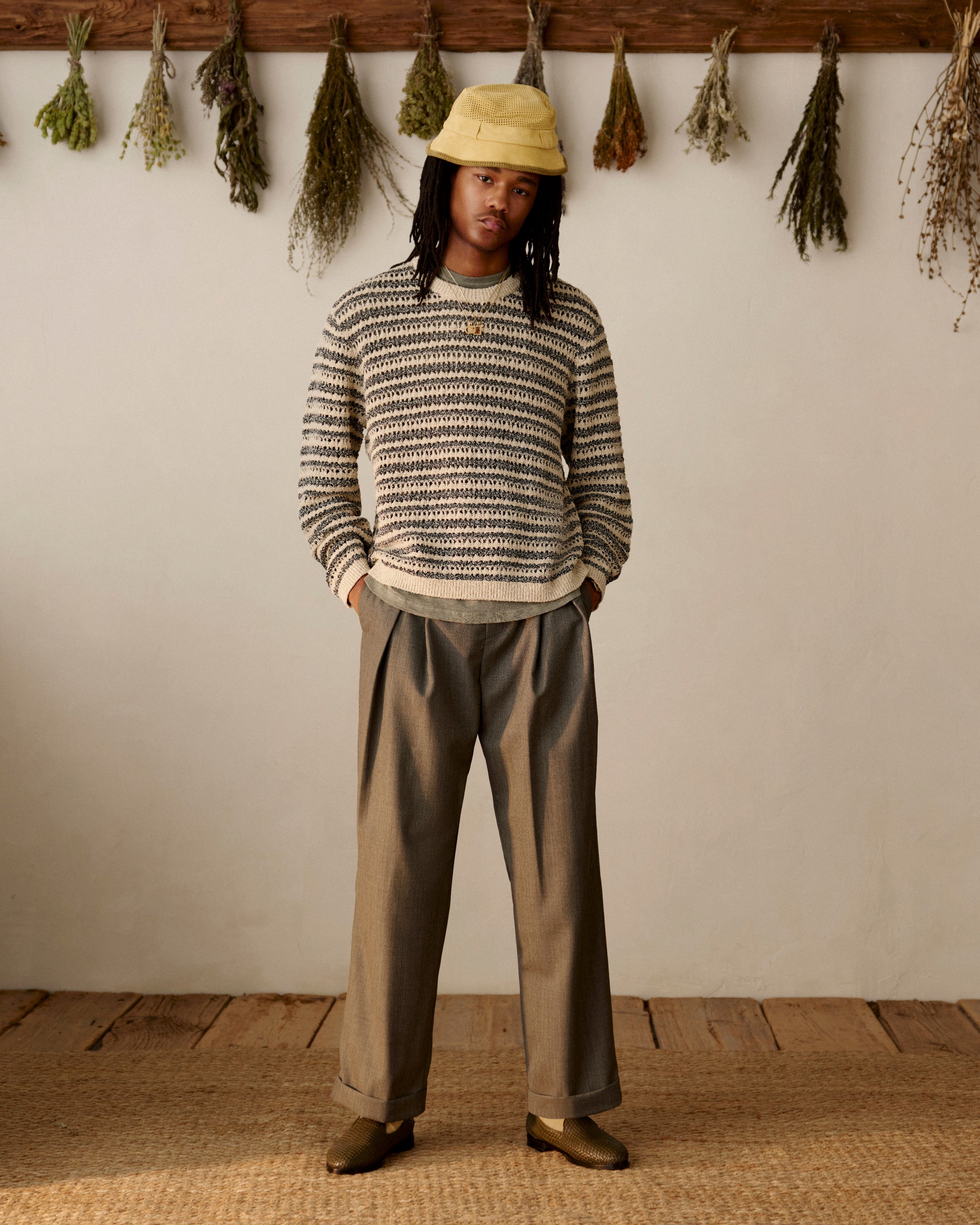 Person in a striped sweater, wide-leg trousers, and Leather Raffia Loafer stands indoors with dried herbs on the wall.
