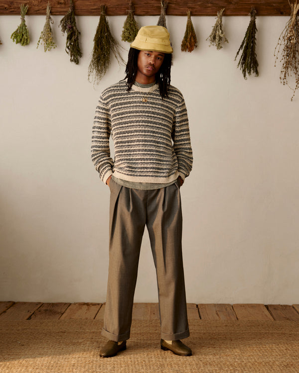 Person in a Bouclé Open Knit Crewneck Sweater, wide-leg pants, and yellow hat stands indoors with dried herbs behind.