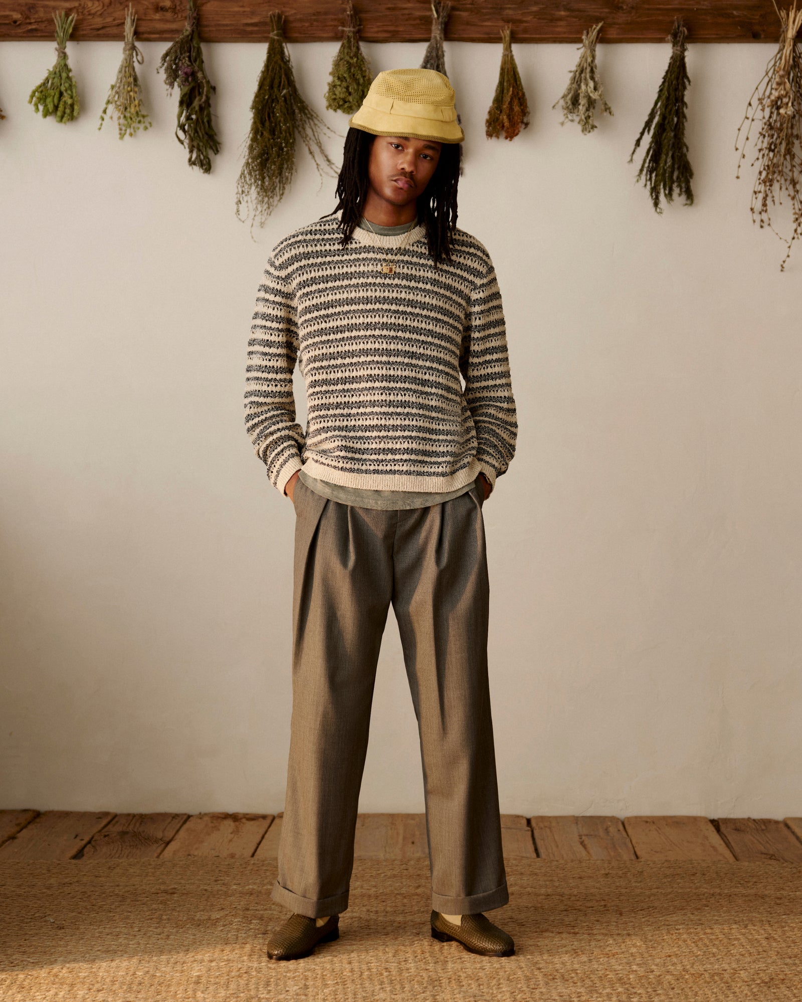 Person wearing a striped sweater, yellow hat, and Lightweight Cuffed Trouser stands indoors with herbs on the wall behind.