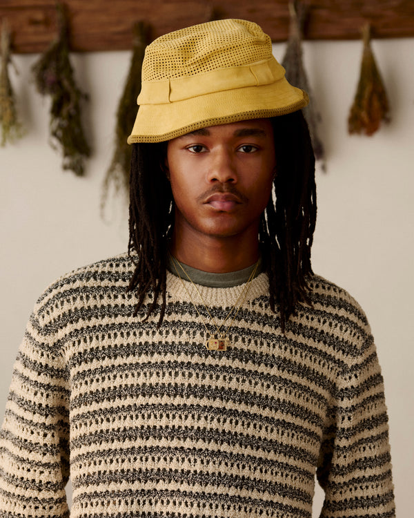 Young person with long dreadlocks wears a Perforated Suede Bucket Hat and striped sweater, standing indoors by hanging herbs.