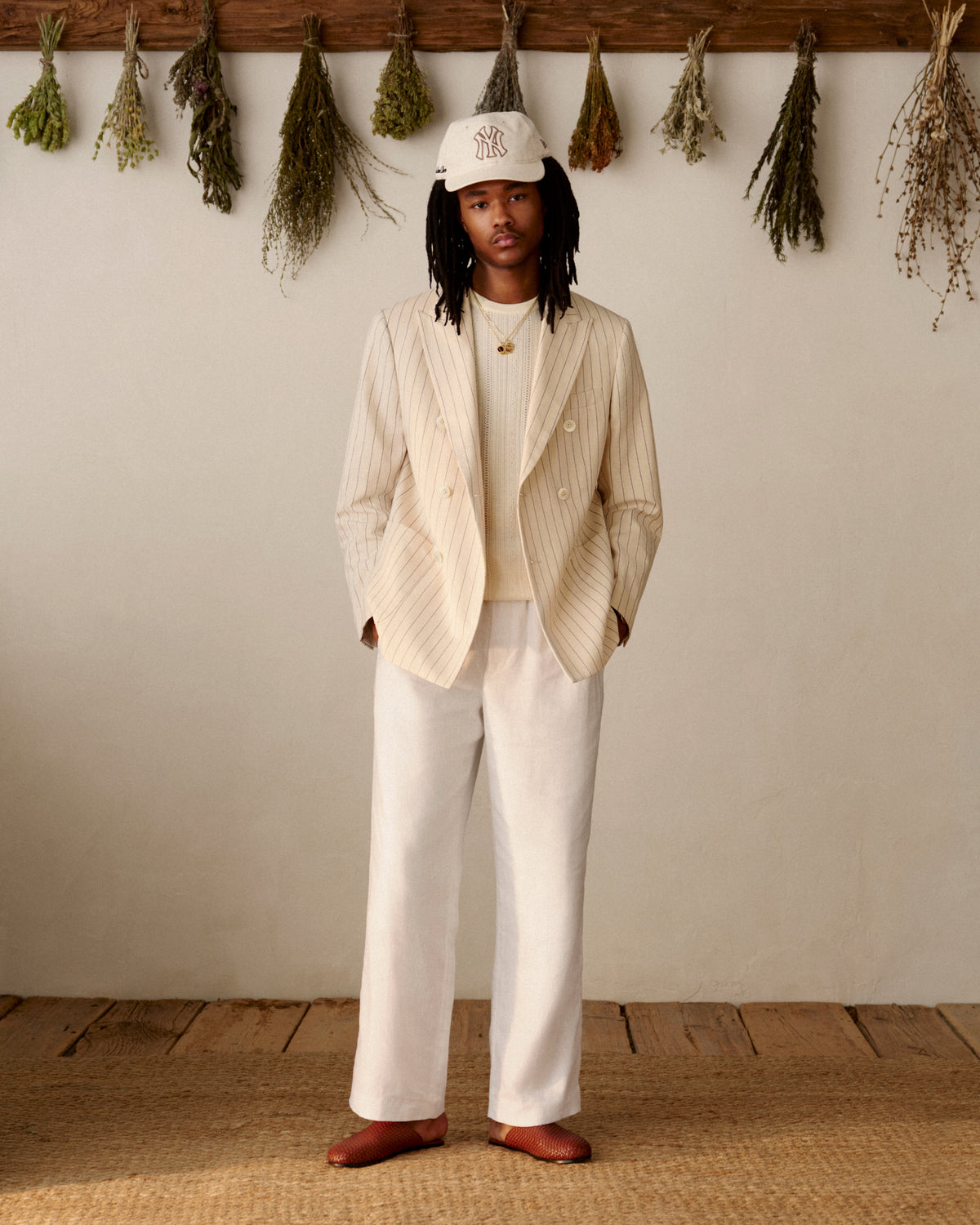 A person in a cream suit and an ALD / New York Yankees Retro Fit Hat stands indoors with dried herbs hanging behind them.