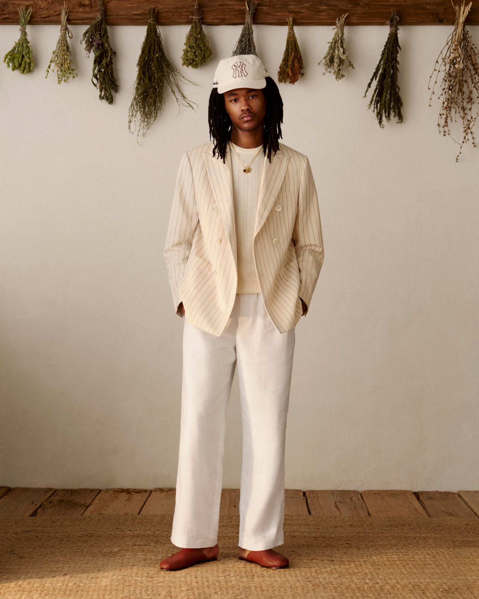 A person in a cream suit and cap stands indoors on a wooden floor, wearing Leather Raffia Mules.