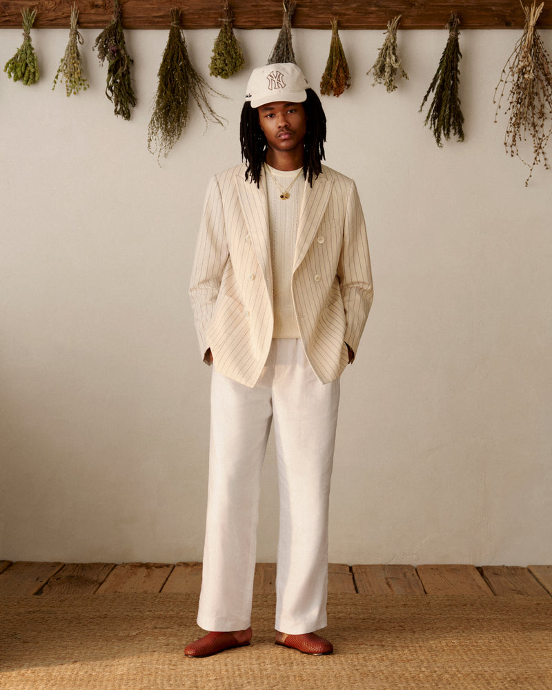 A person in a cream suit and an ALD / New York Yankees Retro Fit Hat stands indoors with dried herbs hanging behind them.
