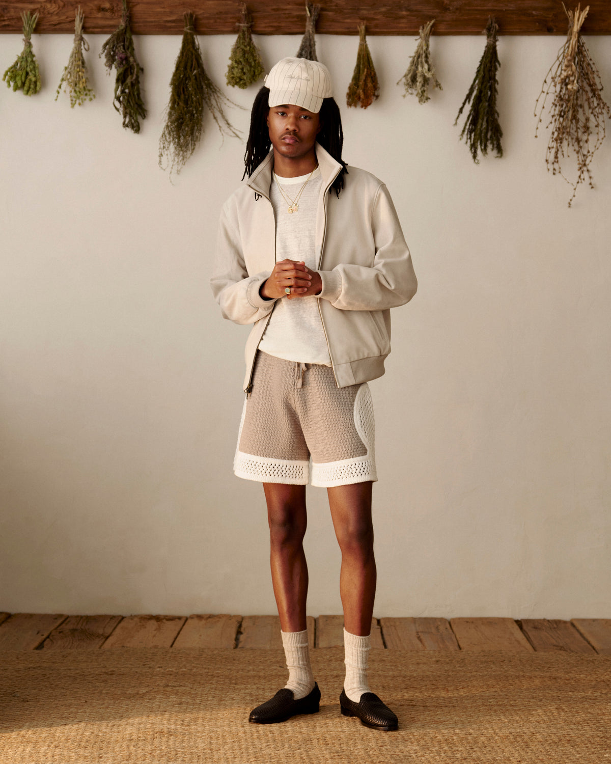 A man in a Suede Track Jacket stands indoors on a woven rug, with dried herbs hanging on the wall behind him.