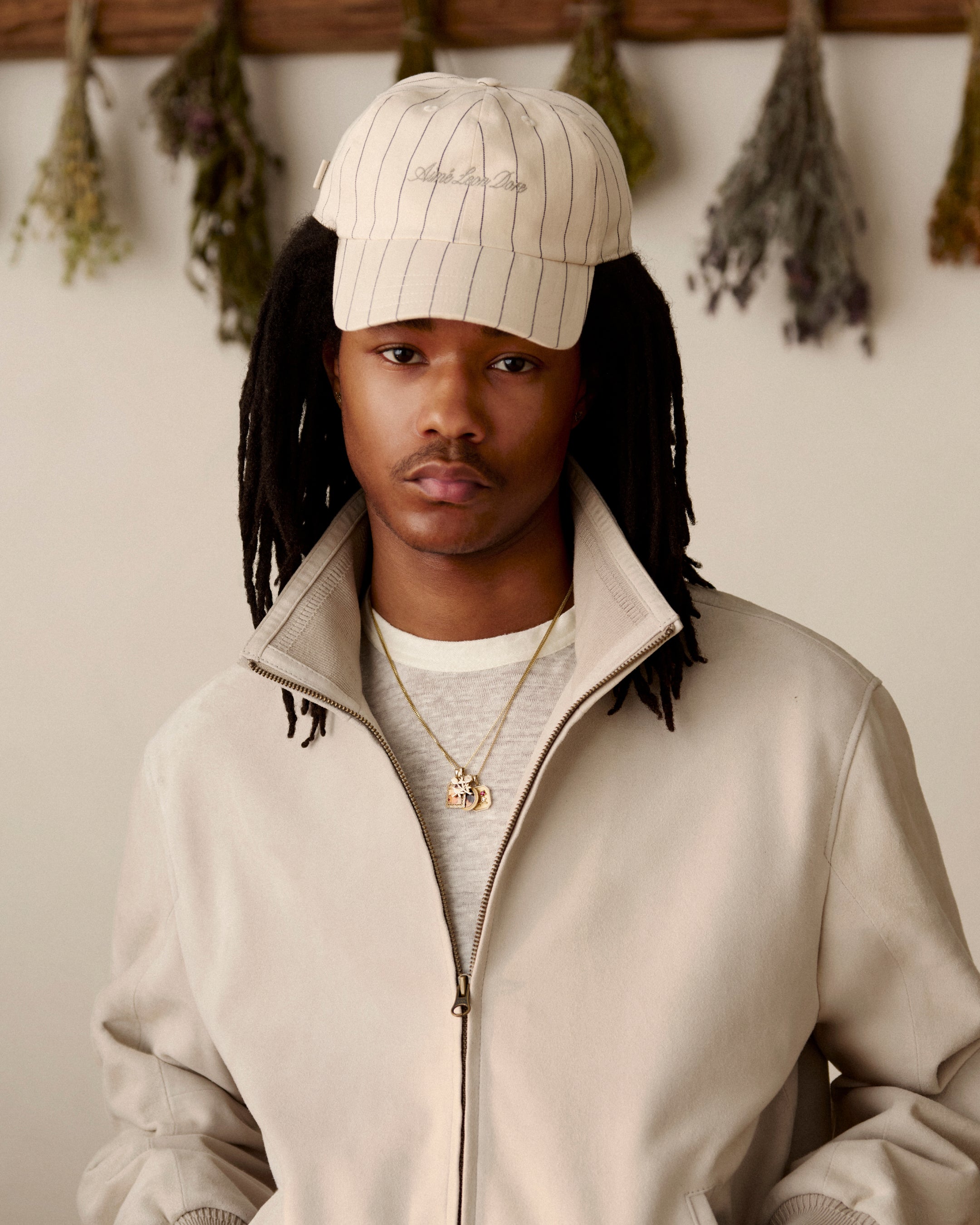 Young man wearing a Pinstripe Suiting Hat and beige jacket, indoors with dried herbs hanging on the wall behind him.
