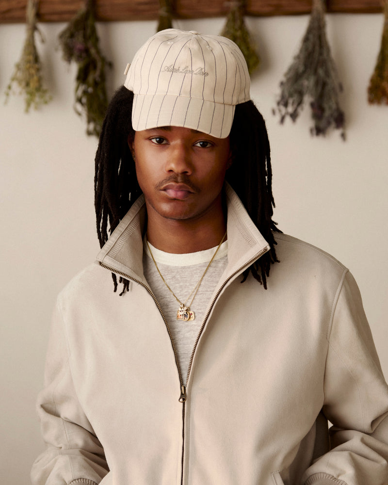 Young man wearing a Pinstripe Suiting Hat and beige jacket, indoors with dried herbs hanging on the wall behind him.