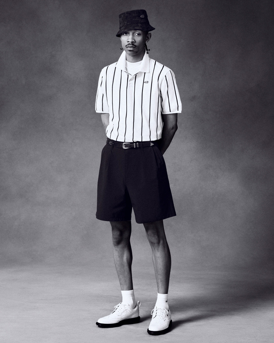 Man in ALD Golf Cordura Double Pleated Short, striped polo, bucket hat, and white shoes stands by gray studio backdrop.