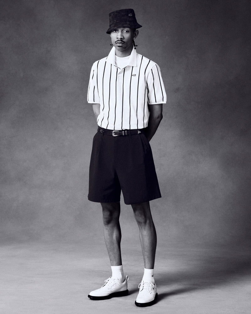 A man in an ALD Golf Polo, black shorts, bucket hat, and white shoes stands against a gray background.
